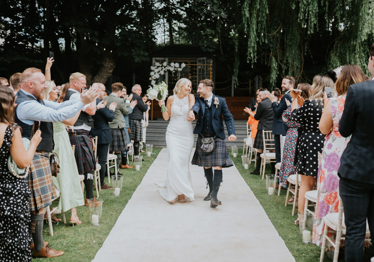 Bride and groom smile together as they walk back down the aisle at The Venue, Eskmills while their guests clap